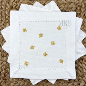 Puede incluir: Servilletas cuadradas blancas con una superficie texturizada y bordado dorado de flor de lis. Las servilletas tienen un borde cosido decorativo y están apiladas sobre una superficie tejida.