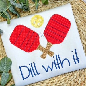 Könnte beinhalten: Weißes T-Shirt mit roten und goldenen gestickten Pickleball-Schlägern und dem Text "Dill with it" in blauer Stickerei.
