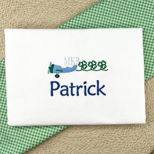 May include: White onesie with blue embroidery of a small airplane flying over a row of green shamrocks. The name "Patrick" is embroidered below the airplane.
