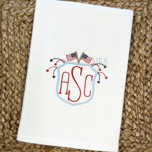 May include: A white embroidered kitchen towel with a blue and red shield design. The shield has the letters "ASC" inside and is topped with two small American flags. Fireworks are coming out of the top of the shield.