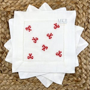 May include: White linen napkins featuring red candy cane embroidery. The square napkins have a stitched border and are stacked. The logo "MKB" is visible in the top right corner.