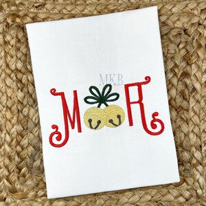 May include: White fabric with red embroidered letters "M&R" and two gold bells. A green bow is embroidered between the letters. The letters have a decorative, curly design. The background is a woven, tan material.
