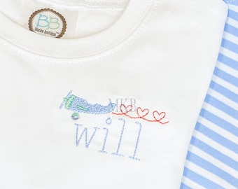 Op de afbeelding: Wit T-shirt met een blauw vliegtuig en rode harten geborduurd erop. De tekst "Will" is geborduurd onder het vliegtuig. Het shirt heeft een bruin label met de tekst "Blanka Boutique" erop.