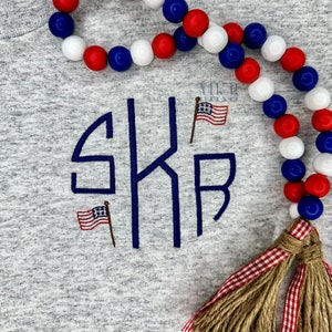 Puede incluir: Una camiseta gris con un monograma bordado en azul "SKB" y dos pequeñas banderas americanas. Una guirnalda de cuentas rojas, blancas y azules con un borla de cuadros rojos y marrones cuelga a un lado.
