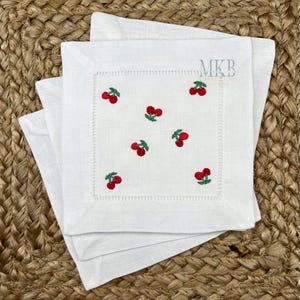 May include: Set of three white linen napkins with embroidered red cherries. The napkins have a white border and the text "MKB" is embroidered in the top right corner.