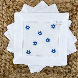 May include: White linen cocktail napkins with blue embroidered Star of David designs. The napkins have a decorative stitched border and the initials "MKB" are visible in the corner. The napkins are stacked on a woven surface.