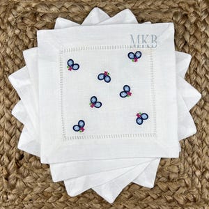 May include: Set of four white linen napkins with a blue and pink embroidered blueberry design. The napkins have a white stitched border.