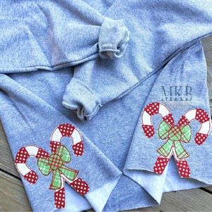 May include: Gray sweatshirt with red and white candy cane and green bow applique on the sleeves. The sweatshirt has a split on the side seams. The text "MKR MIX" is visible on the sweatshirt.