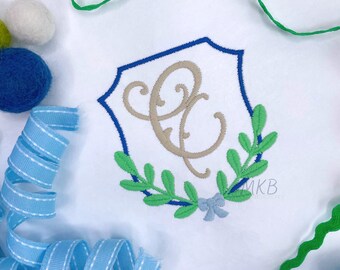 May include: A white fabric with a blue and green embroidered monogram. The monogram is a stylized letter "C" inside a shield with a wreath of leaves around it. The wreath has a blue bow at the bottom.