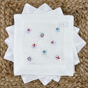 May include: White linen cocktail napkins with embroidered colorful floral and geometric designs. The napkins are square and stacked, with the initials "MKB" embroidered in the upper right corner. The napkins are on a woven surface.