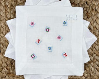 May include: White linen cocktail napkins with embroidered colourful floral and geometric designs. The napkins are square and stacked, with the initials "MKB" embroidered in the upper right corner. The napkins are on a woven surface.