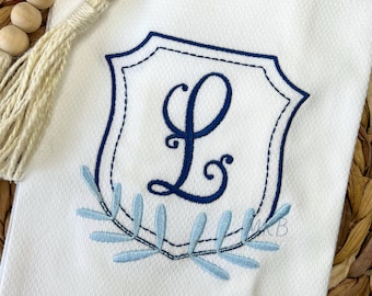 May include: A white kitchen towel with a blue embroidered crest featuring the letter L and a blue leafy design.