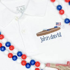 May include: White polo shirt with a brown boat and American flag embroidery. The text "john david" is embroidered below the boat. A red, white, and blue beaded necklace is in the foreground.