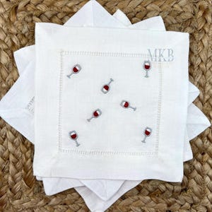 May include: White linen cocktail napkins with embroidered red wine glasses. The napkins have a stitched border and are stacked on a woven surface. The logo "MKB Embroidery Designs" is visible in the upper right corner.