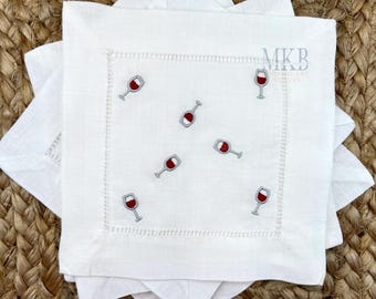 Peut inclure: Serviettes de cocktail en lin blanc avec des verres de vin rouge brodés. Les serviettes ont une bordure cousue et sont empilées sur une surface tissée. Le logo "MKB Embroidery Designs" est visible en haut à droite.