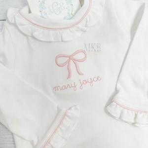 May include: White long-sleeve shirt with a pink bow and the name "mary joyce" embroidered on the front. The shirt has a ruffled collar and cuffs.
