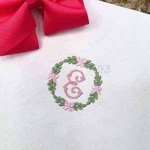 May include: A white fabric with a pink and green embroidered wreath featuring the letter E in the centre. The wreath is made of leaves and flowers.