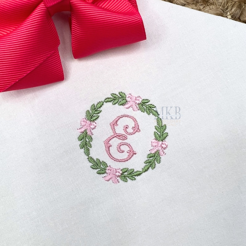 May include: A white fabric with a pink and green embroidered wreath featuring the letter E in the centre. The wreath is made of leaves and flowers.