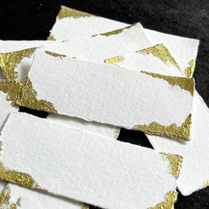Puede incluir: Un conjunto de etiquetas rectangulares de papel blanco con bordes rasgados, cada una adornada con detalles de pan de oro en las esquinas. Las etiquetas están dispersas sobre un fondo oscuro, creando un contraste.