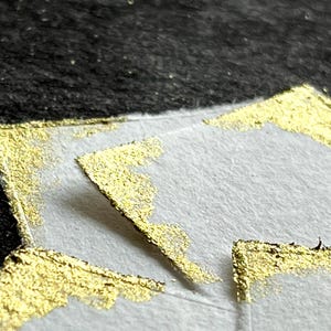 Könnte beinhalten: Eine weiße Papieroberfläche mit strukturierten Blattgoldakzenten an den Ecken. Das Blattgold hat eine metallisch reflektierende Oberfläche mit unregelmäßigen, gezackten Kanten, die einen Kontrast zum matten weißen Papier und dem dunklen Hintergrund bilden.