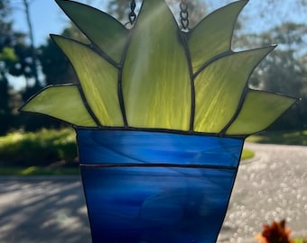 May include: A stained glass succulent plant in a blue pot hanging from a chain. The plant has green leaves and the pot has a blue and white swirl pattern.