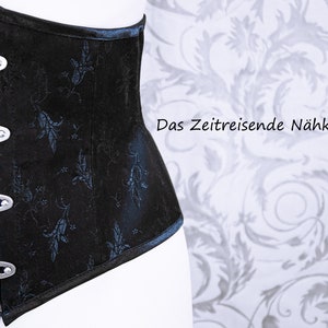 May include: Black corset with silver clasps and a floral pattern. The corset has a structured design and a slight curve. The text "Das Zeitreisende N&auml;hk&auml;stchen" is visible in the background. The corset is a fashion item.