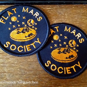 Könnte beinhalten: Zwei schwarze und orangefarbene gestickte Patches mit dem Text "Flat Mars Society" auf jedem. Die Patches zeigen ein stilisiertes Bild einer flachen Erde mit Sternen und Planeten drumherum.