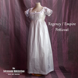 Könnte beinhalten: Ein weißes Regency/Empire Petticoat-Kleid auf einer Schaufensterpuppe. Das Kleid hat einen quadratischen Ausschnitt, kurze Ärmel und ein eng anliegendes Oberteil mit Schnürdetail. Der Rock ist lang und fließend. Der Text auf dem Bild lautet "Regency / Empire Petticoat" und "Das Zeitreisende Nähkästchen".