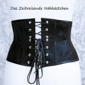 May include: Black corset with a floral pattern and a lace-up back. The corset has silver eyelets and black lacing. The text "Das Zeitreisende N&auml;hk&auml;stchen" is at the top. The corset is displayed on a white mannequin.