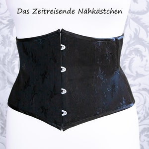 May include: Black corset with a floral pattern and three hook-and-eye closures. The corset is displayed on a mannequin. The text "Das Zeitreisende N&auml;hk&auml;stchen" is at the top.