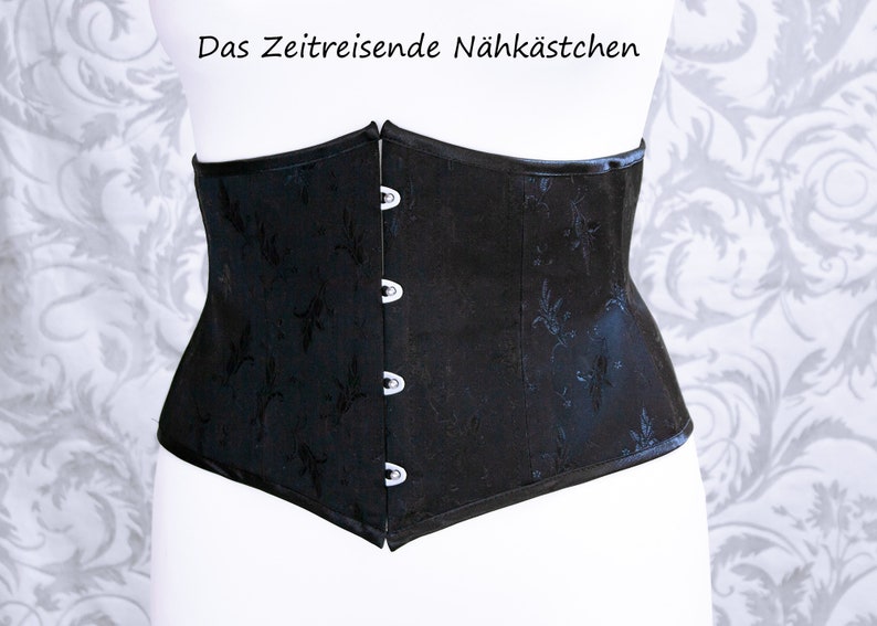 May include: Black corset with a floral pattern and three hook-and-eye closures. The corset is displayed on a mannequin. The text "Das Zeitreisende N&auml;hk&auml;stchen" is at the top.
