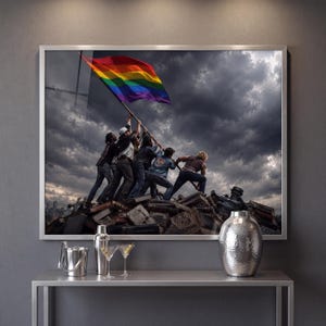 May include: A framed wall art print depicting a group of people raising a rainbow pride flag atop a pile of books and debris under a dark, cloudy sky. One book is clearly visible with the text "EQUALITY IS HUMAN" printed on its cover.