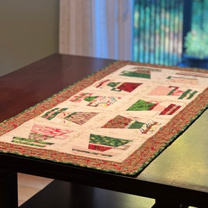 May include: A rectangular quilted table runner featuring a repeating pattern of stylized teacups in red and green on a cream background. The border is a braided design in red, green, and gold. The runner is placed on a dark brown table.