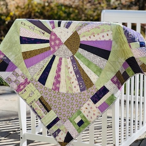 May include: A handmade quilt with a sunburst design in shades of green, purple, and brown. The quilt is draped over a white crib, showcasing its intricate patchwork and geometric patterns. The quilt's edges are finished with a decorative stitch.