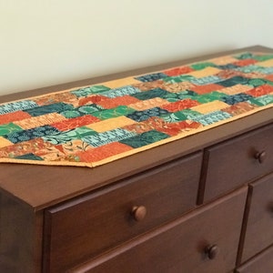 May include: A colorful patchwork table runner with a geometric design. The runner features a variety of fabric patterns in shades of orange, green, blue, and yellow. It is placed on a dark brown wooden dresser.