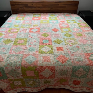 May include: A patchwork quilt with a variety of floral patterns in shades of pink, green, and white. The quilt is laid out on a bed with a wooden headboard.