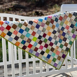 May include: A colorful patchwork quilt with a green border, hanging on a white picket fence. The quilt is made of squares of different fabrics, each with a unique pattern.