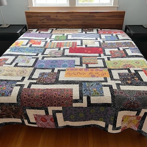 May include: A white quilt with a black geometric pattern. The quilt is made of different fabrics with various colorful patterns. The quilt is on a bed with two black nightstands.