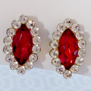May include: A pair of red and clear rhinestone earrings. The earrings are shaped like a marquise and have a silver tone setting.