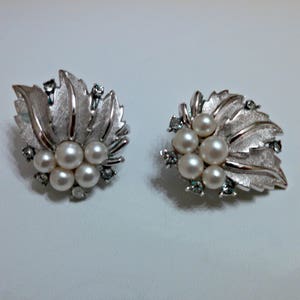 May include: A pair of silver earrings with a leaf design and pearl accents. The leaves are textured and have a slightly raised design. The pearls are white and are clustered together in the center of each earring.