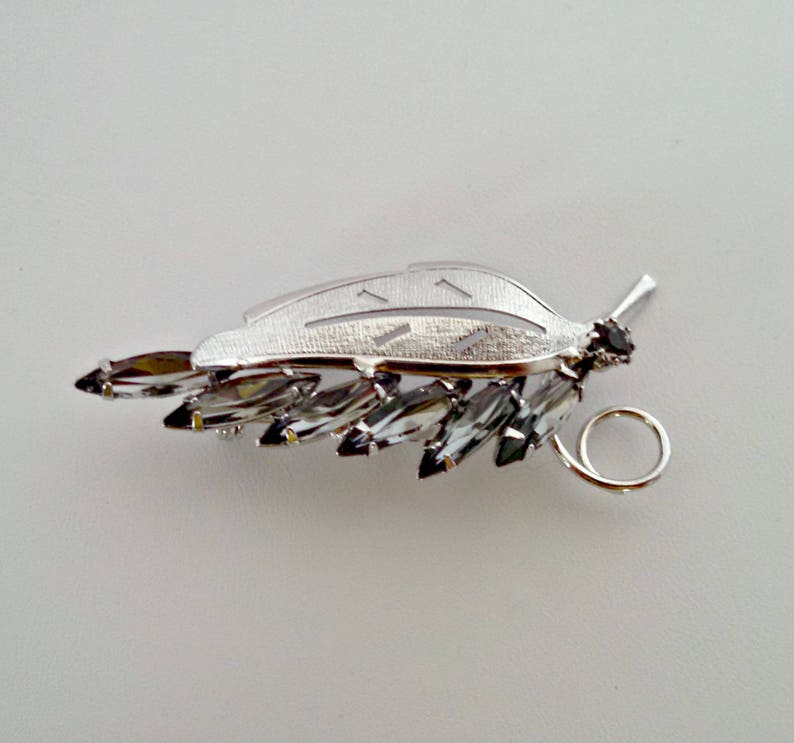 May include: A silver leaf brooch with a cluster of clear and black rhinestones. The brooch has a silver loop on the back for attaching to clothing.