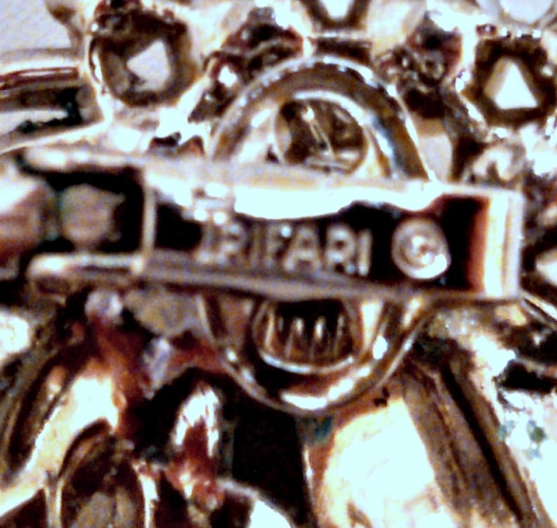 May include: Close-up of a silver-toned metal brooch with a floral design. The back of the brooch has the word "TRIFARI" and a copyright symbol.