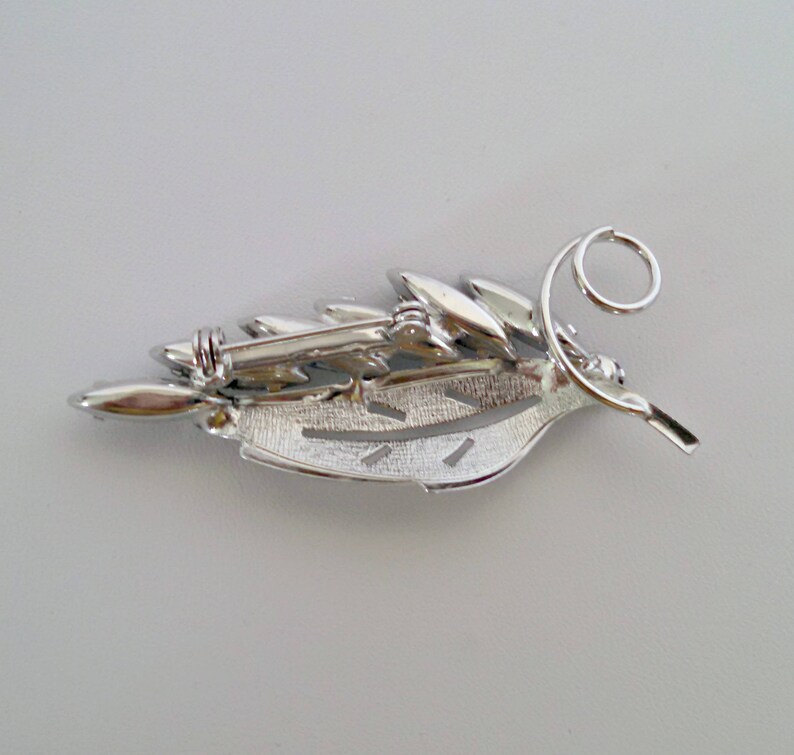 May include: Silver tone brooch in the shape of a leaf with clear rhinestones. The brooch has a pin and a safety catch on the back.