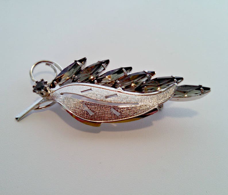 May include: A silver tone brooch in the shape of a leaf with a textured surface. The leaf is adorned with dark gray rhinestones.
