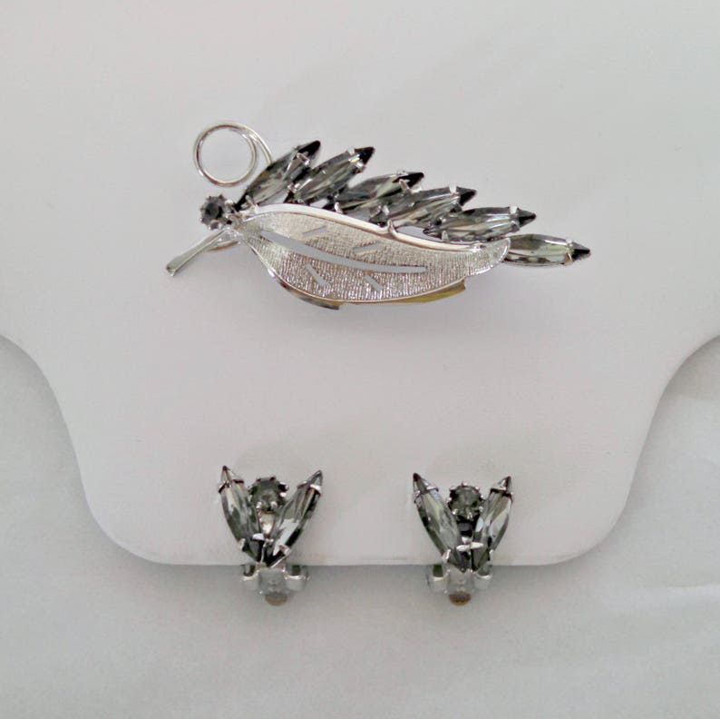 May include: A silver leaf-shaped brooch with clear rhinestones and a matching pair of clip-on earrings with clear rhinestones.