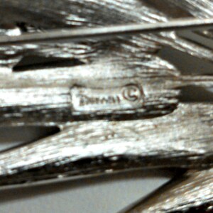 May include: Close-up of a silver-toned brooch with a textured design. The brooch has a small, circular, raised inscription that reads "Tuscany (C)".