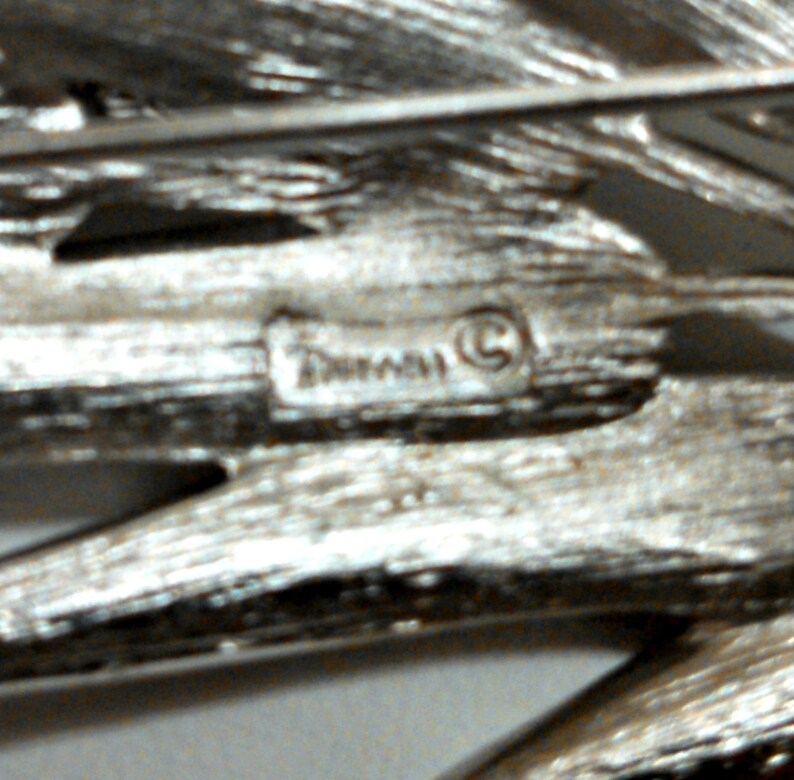 May include: Close-up of a silver-toned brooch with a textured design. The brooch has a small, circular, raised inscription that reads "Tuscany (C)".