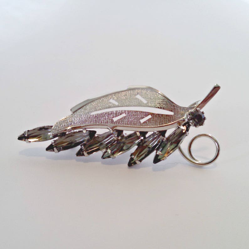 May include: A silver leaf-shaped brooch with a row of dark gray rhinestones. The brooch has a silver loop on the back for attaching to clothing.