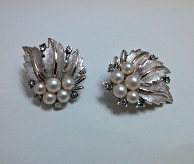 May include: A pair of silver earrings with a leaf design and pearl accents. The leaves are textured and have a slightly raised design. The pearls are white and are clustered together in the center of each earring.