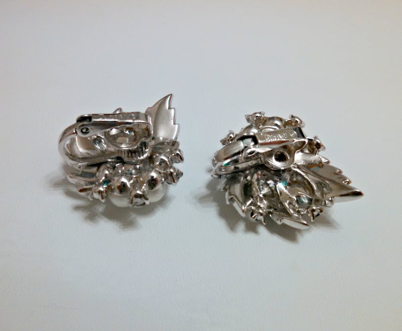 May include: A pair of silver tone clip-on earrings with a floral design and clear rhinestones. The earrings have a silver tone metal back.